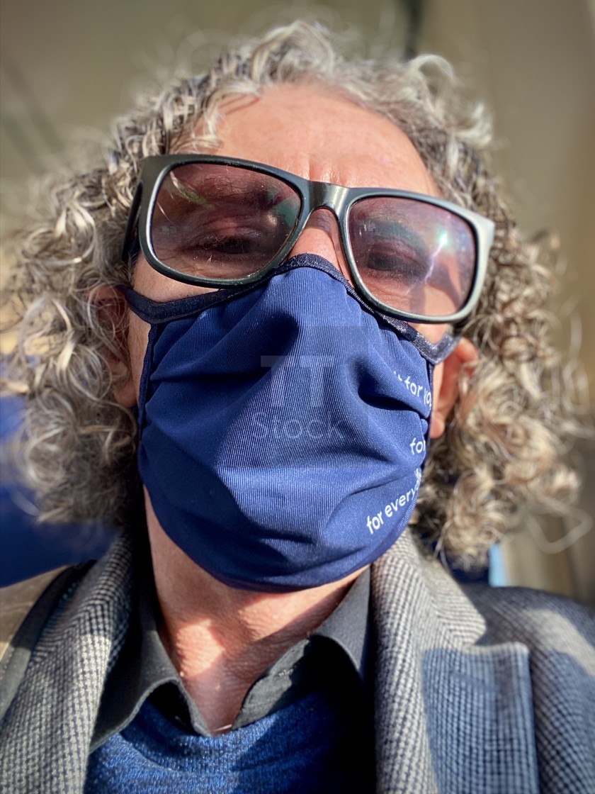 "Mature older caucasian man with grey curly hair wearing sunglasses and blue face mask for protection against COVID-19 corona virus spreading, traveling alone on the NS commuter train. Golden sunlight shines through carriage window. Train travel journey co" stock image