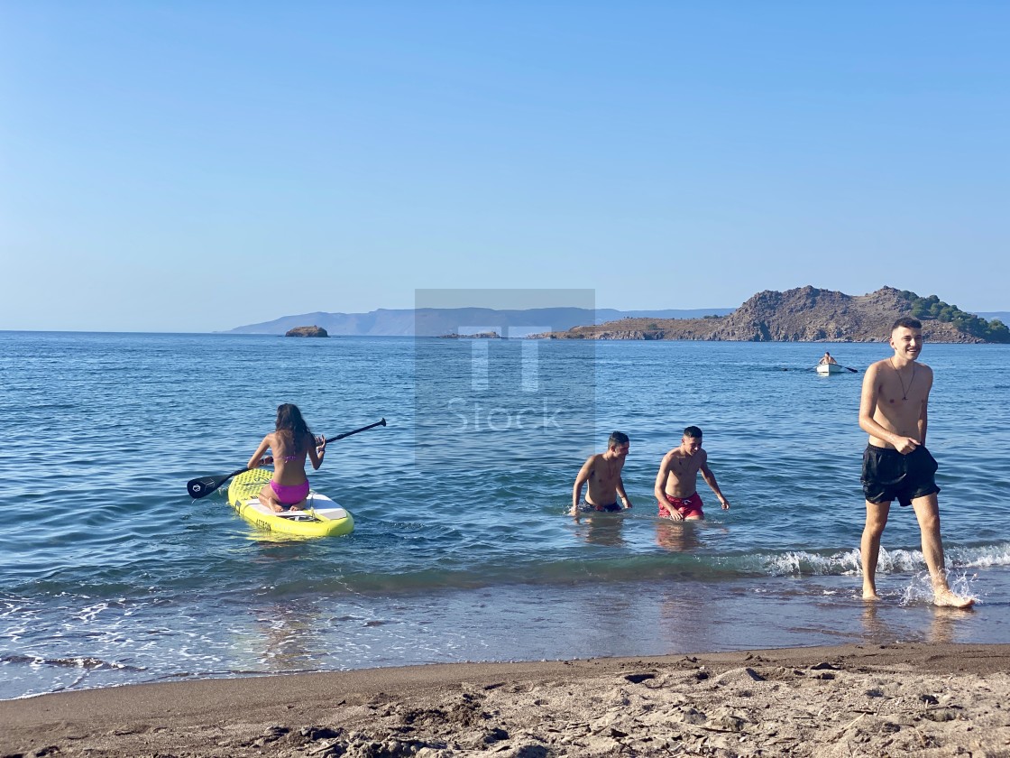 "Young woman wearing a pink bikini on the beach kneeling on sup board, youths walk out the sea." stock image