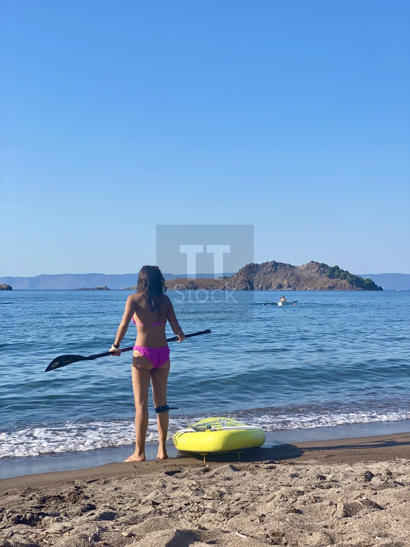 "Young woman wearing a pink bikini standing on the beach with a yellow sup board attached." stock image