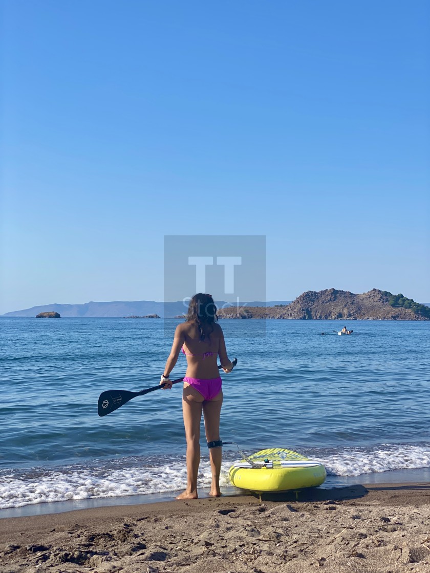 "Young woman wearing a pink bikini standing on the beach with a yellow sup board attached." stock image