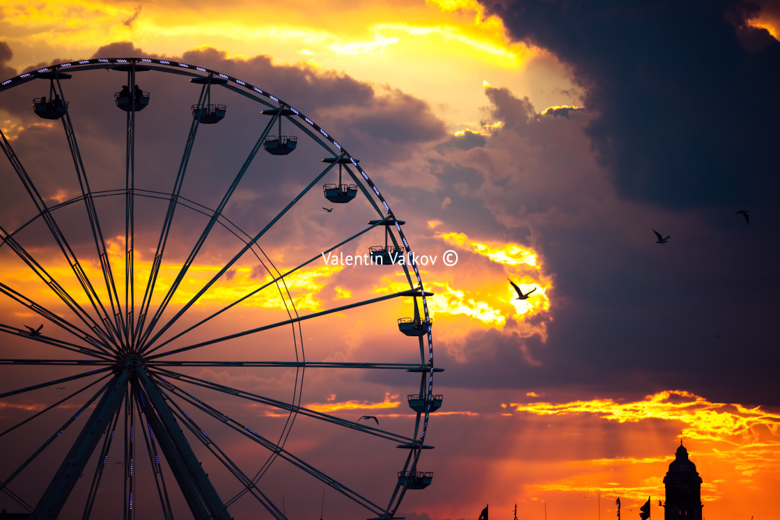 "Ferris Wheel and illuminations in amusement park during scenic sunset with dramatic sky clouds and flying birds" stock image