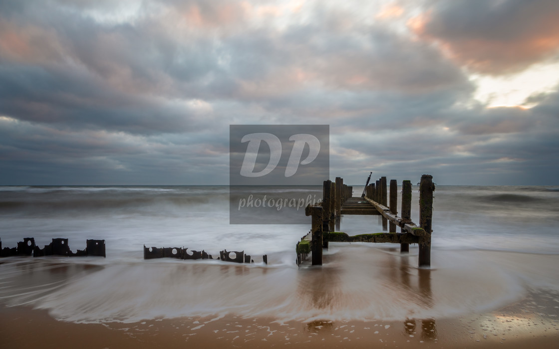 "Image taken on Happisburgh beach Norfolk showing the waves swirling round the sea defences and the light of the rising sun catching the underside of the clouds." stock image