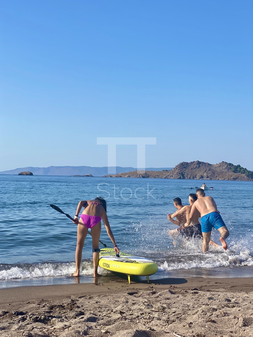 "Young woman wearing a pink bikini on the beach with a sup board, youths jump in the sea." by TTStock - $7.54 "Young woman wearing a pink bikini on the beach with a sup board, youths jump in the sea." stock image