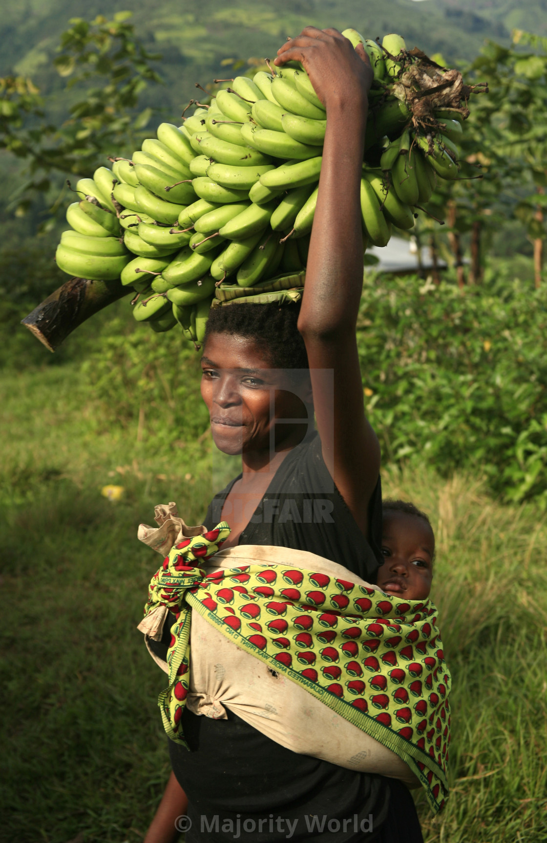 "African woman with baby on her back and green bananas on her head in..." stock image