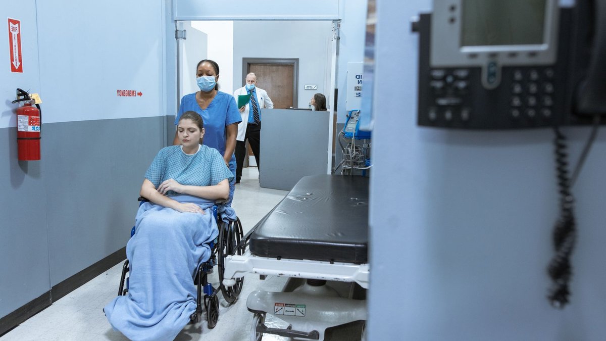 A nurse helps a patient in a wheelchair down a hospital corridor, reflecting care and medical professionalism.
