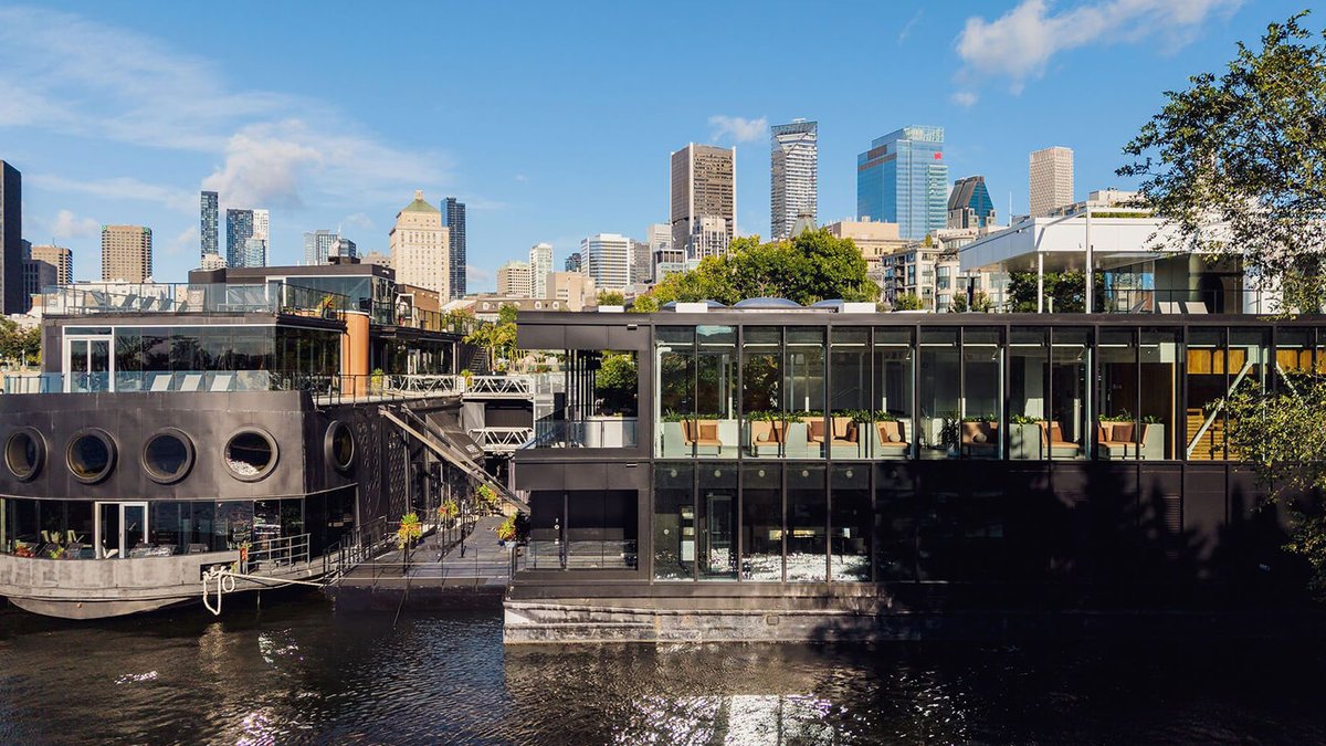 Montreal's Bota Bota Expands with Social Sauna Pavilion Modern black bota bota Pavillon on a waterfront with large windows, outdoor seating, and city skyscrapers visible in the back
