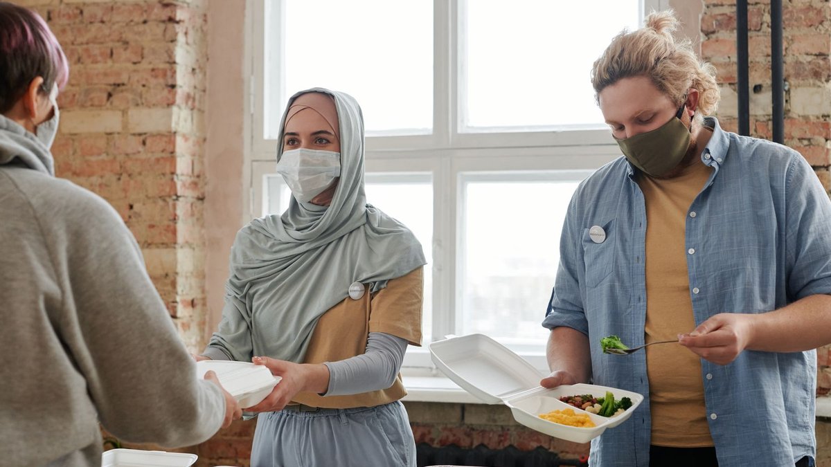 Volunteers wearing masks distribute meals indoors, promoting safety and care.