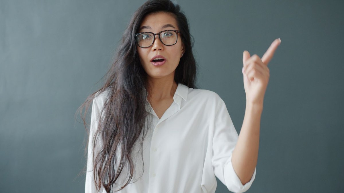 A surprised woman with glasses points upwards against a neutral background, wearing a white blouse.