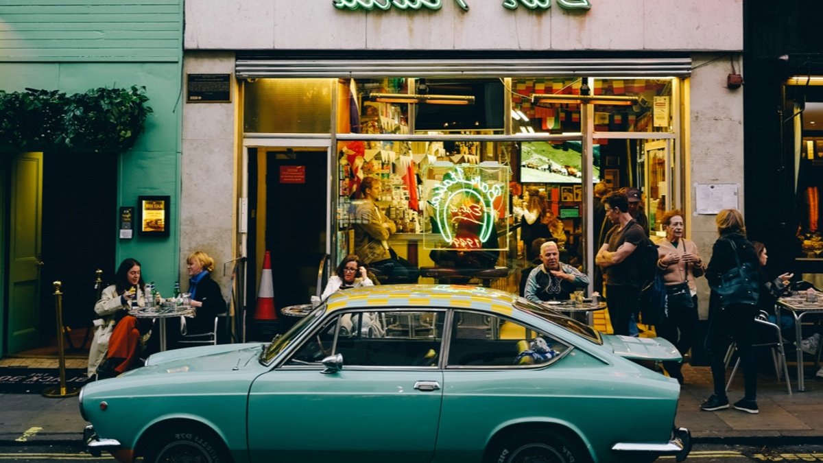 A teal vintage car parked in front of Bar Italia in Soho, with people on the sidewalk and inside the cafe.