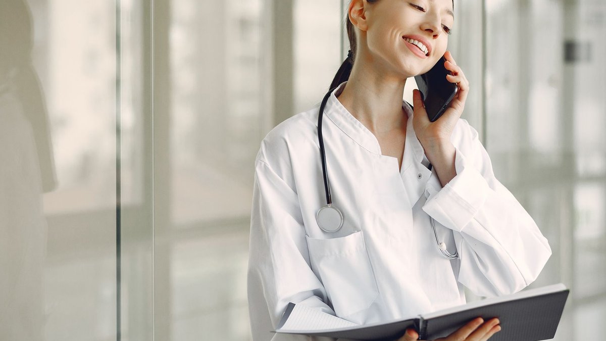 Smiling ethnic female doctor in medical uniform with stethoscope and notebook talking on smartphone while standing in glass h