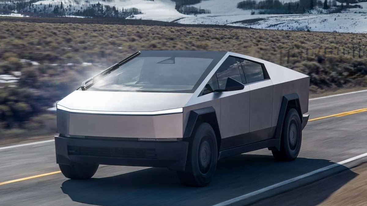 Silver Tesla Cybertruck driving on a paved road with snow-covered hills in the background.