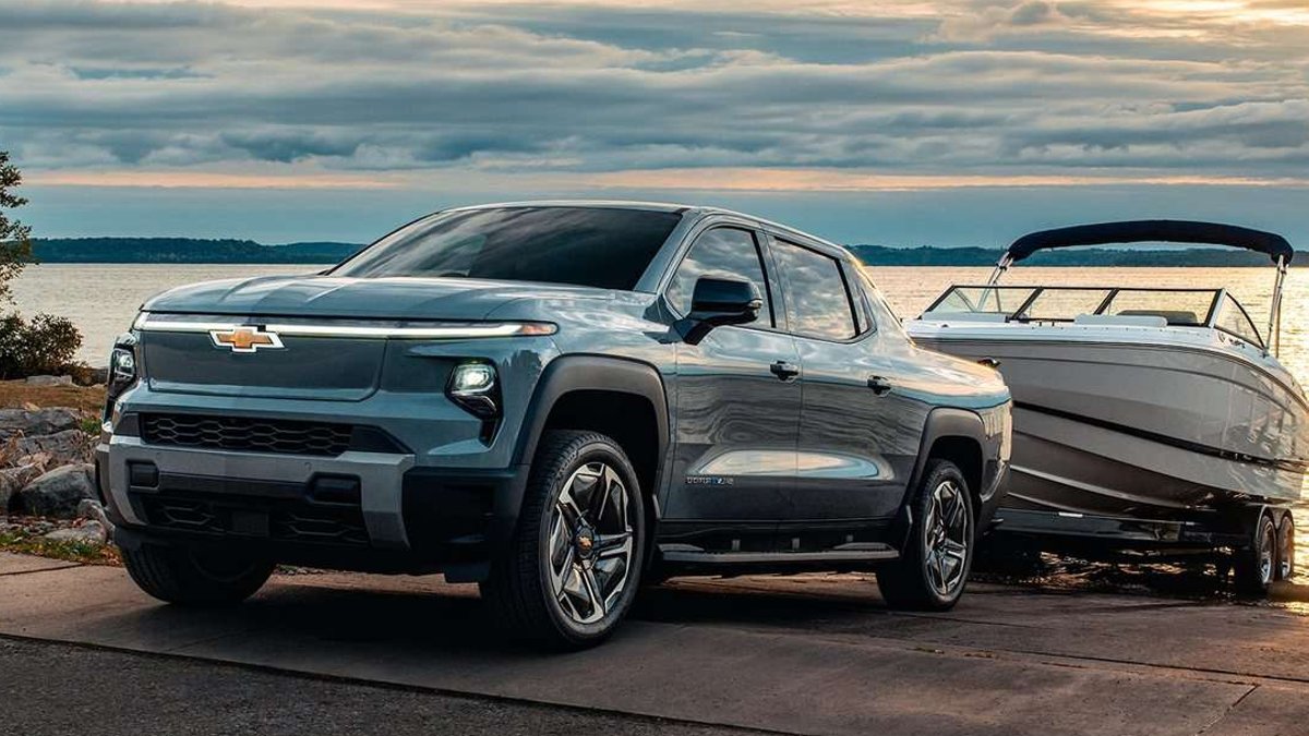 Blue Chevrolet Silverado EV towing a boat on a trailer at a lakeside launch ramp.