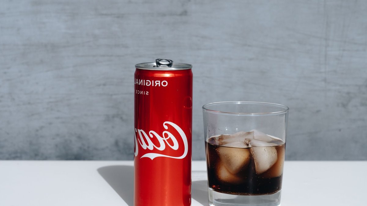 Chilled Coca-Cola can and drink with ice cubes in glass on minimalistic surface.