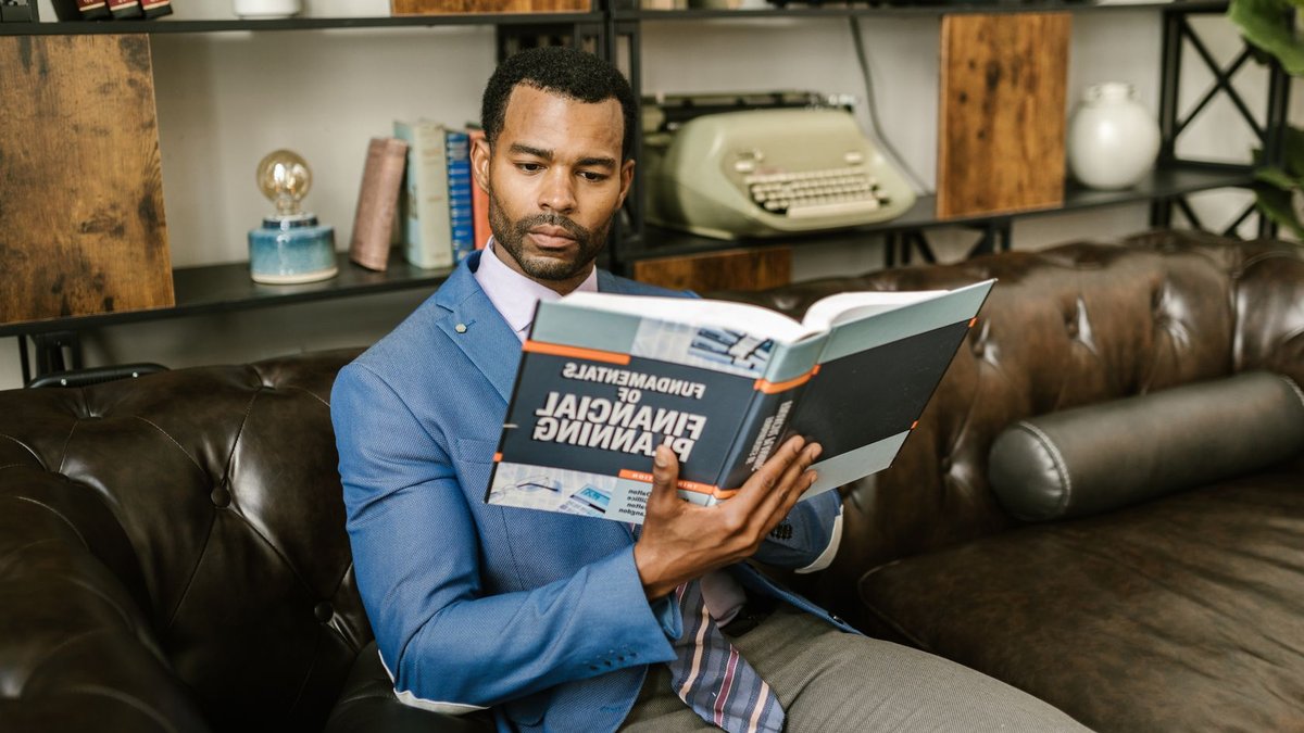 A man in a blue blazer sits on a leather couch reading a financial planning book.