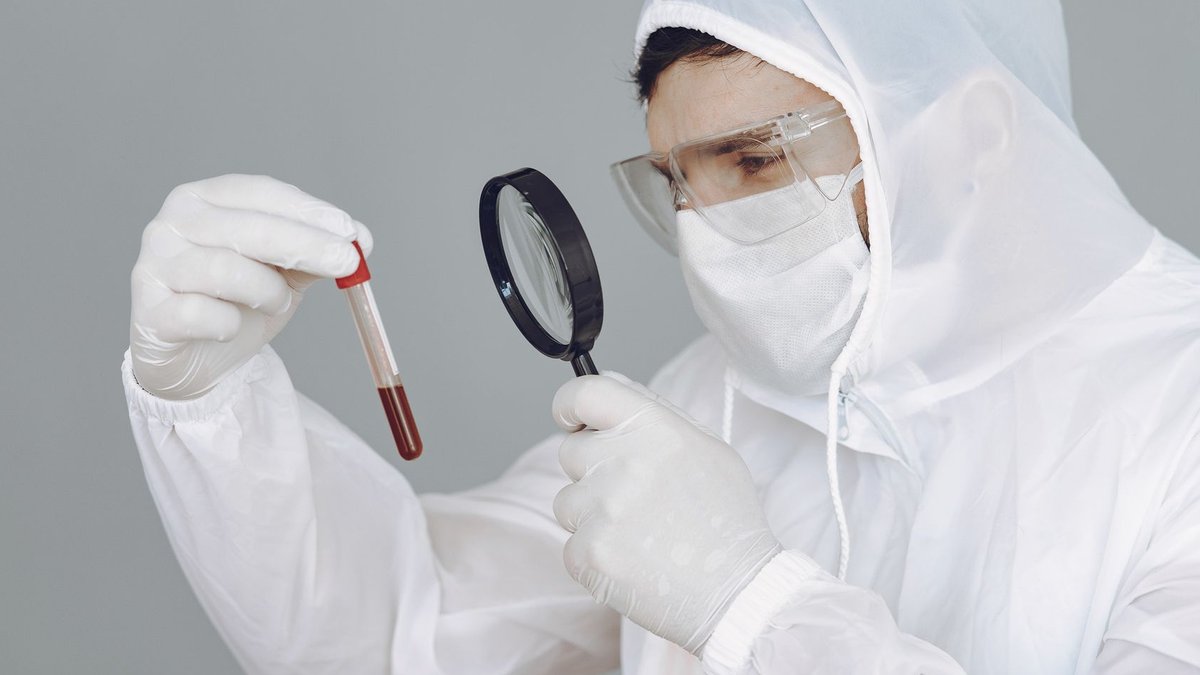 A scientist in protective gear examines a blood sample through a magnifying glass.