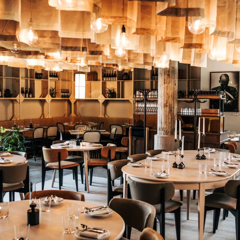 A dining area with round tables set for meals, chairs, shelving with bottles, and unique lighting.
