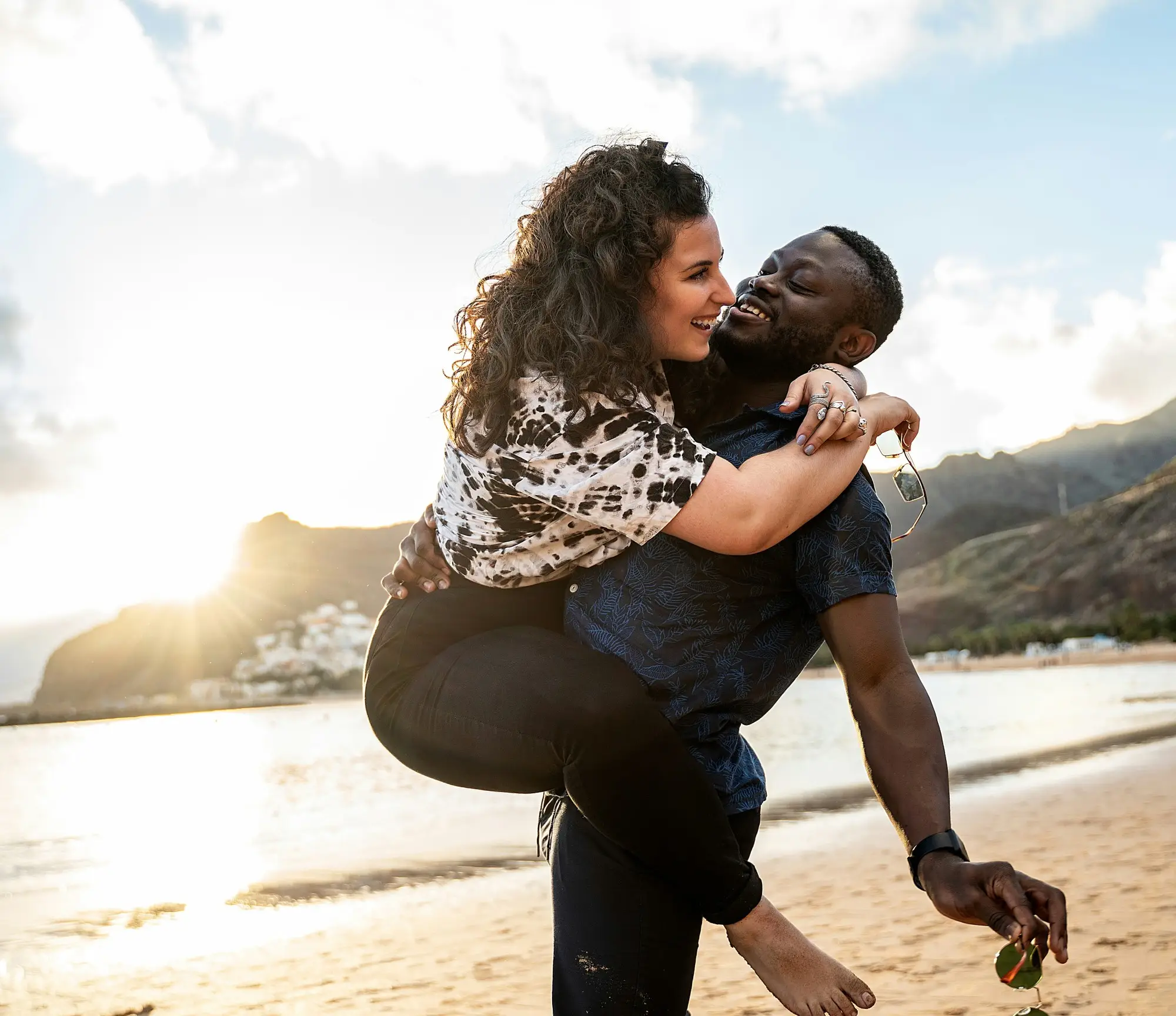 The image depicts two individuals on a beach, with one carrying the other on their back, facing a mountainous landscape during what appears to be sunset.