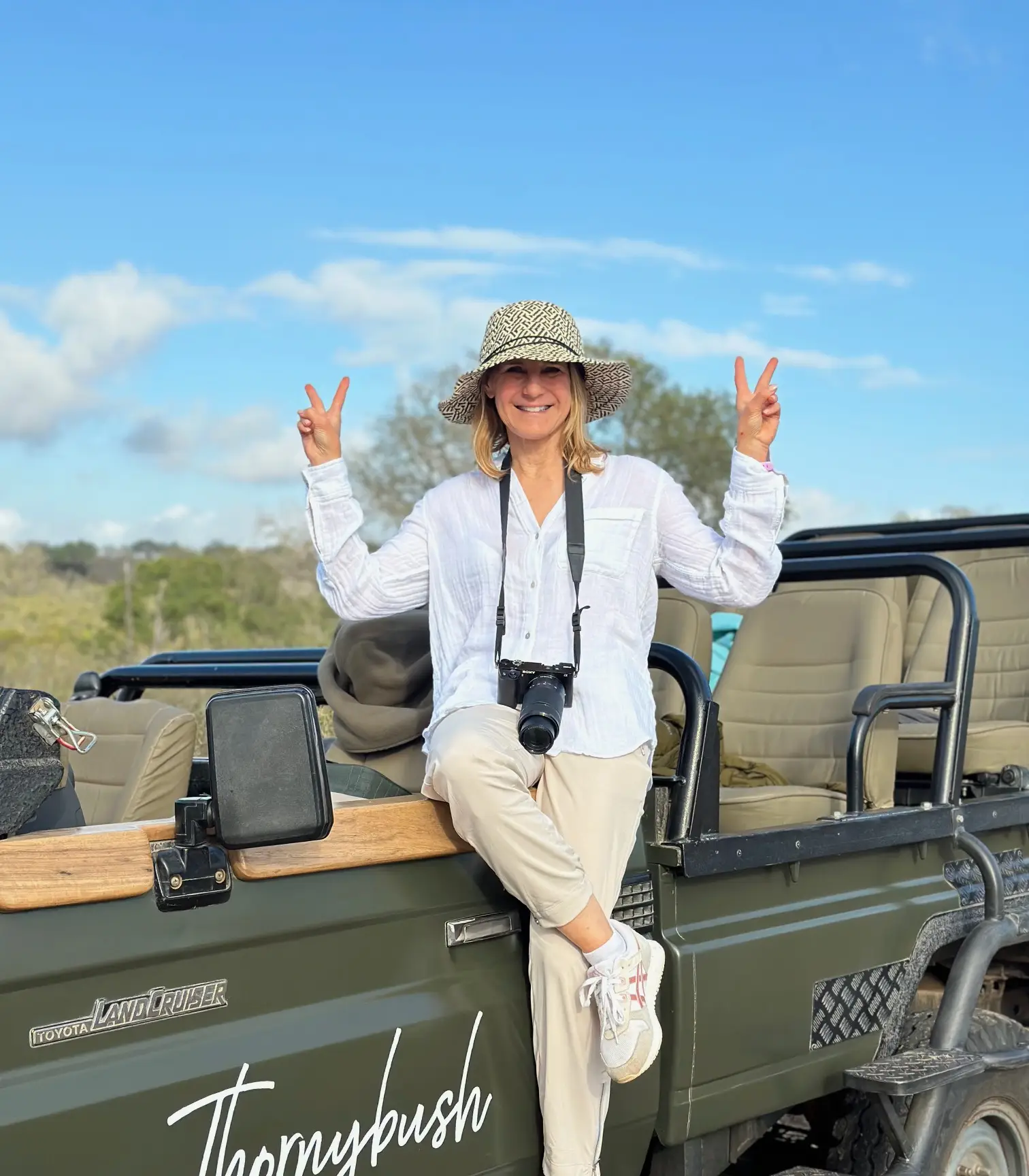 Lee Newell in a white shirt and pants and sun hat with a camera around her neck sitting on the edge of a Jeep that says "Thornybush," on her trip to see some of the best places to visit in South Africa.