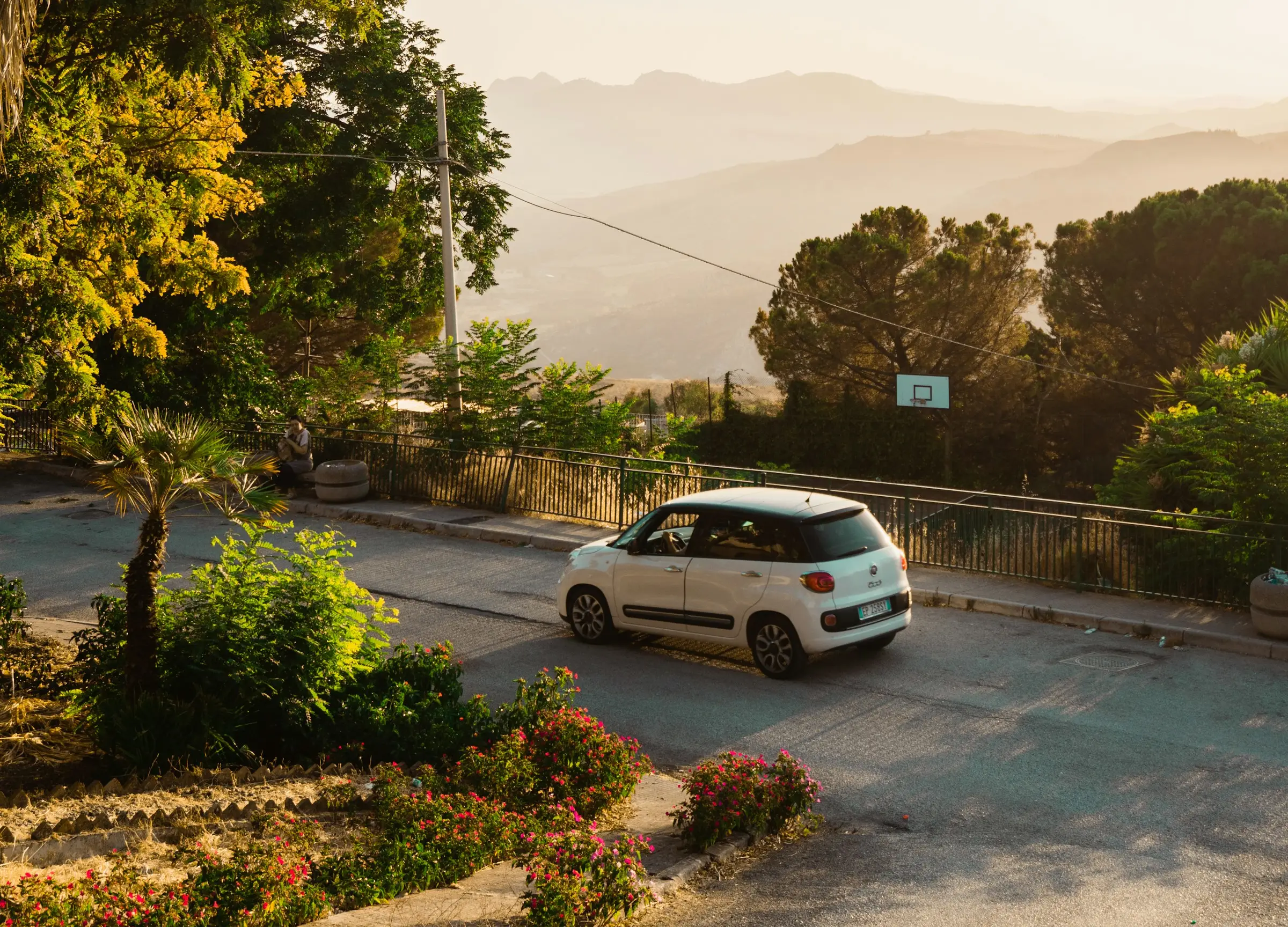 A white car on the road surrounded by foliage with low mountains in the background.