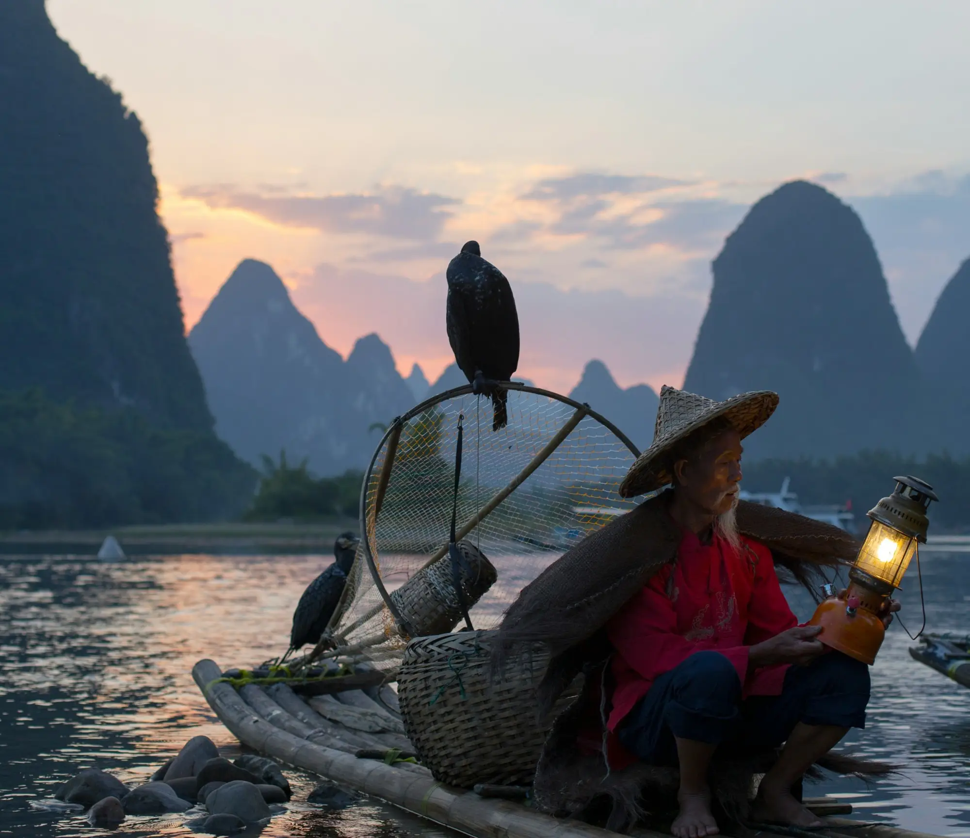 A man sits on a small handmade raft with a lantern at sunset, two birds are perched on the raft with him and there are large rock formations in the ocean ahead of them.