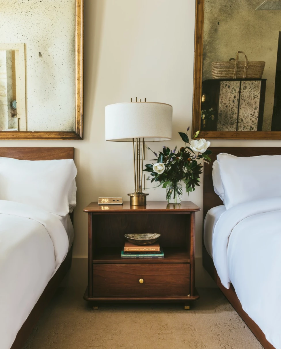 a small wooden table with a bouquet of lowers in between two white beds