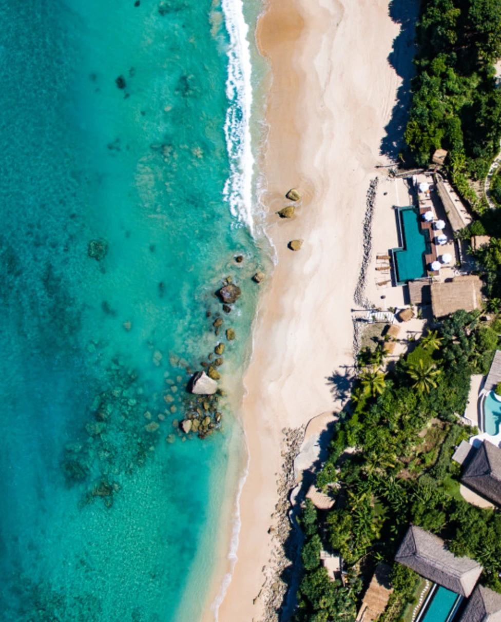 Aerial view of a beautiful beach with a resort nestled in lush jungle with multiple buildings and pools.