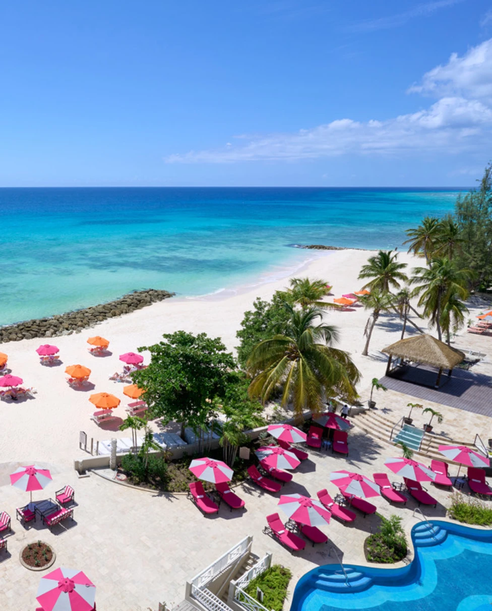 An aerial view of a white sand beach with turquoise wter, pink and orange umbrellas and sun loungers and part of a swimming pool.
