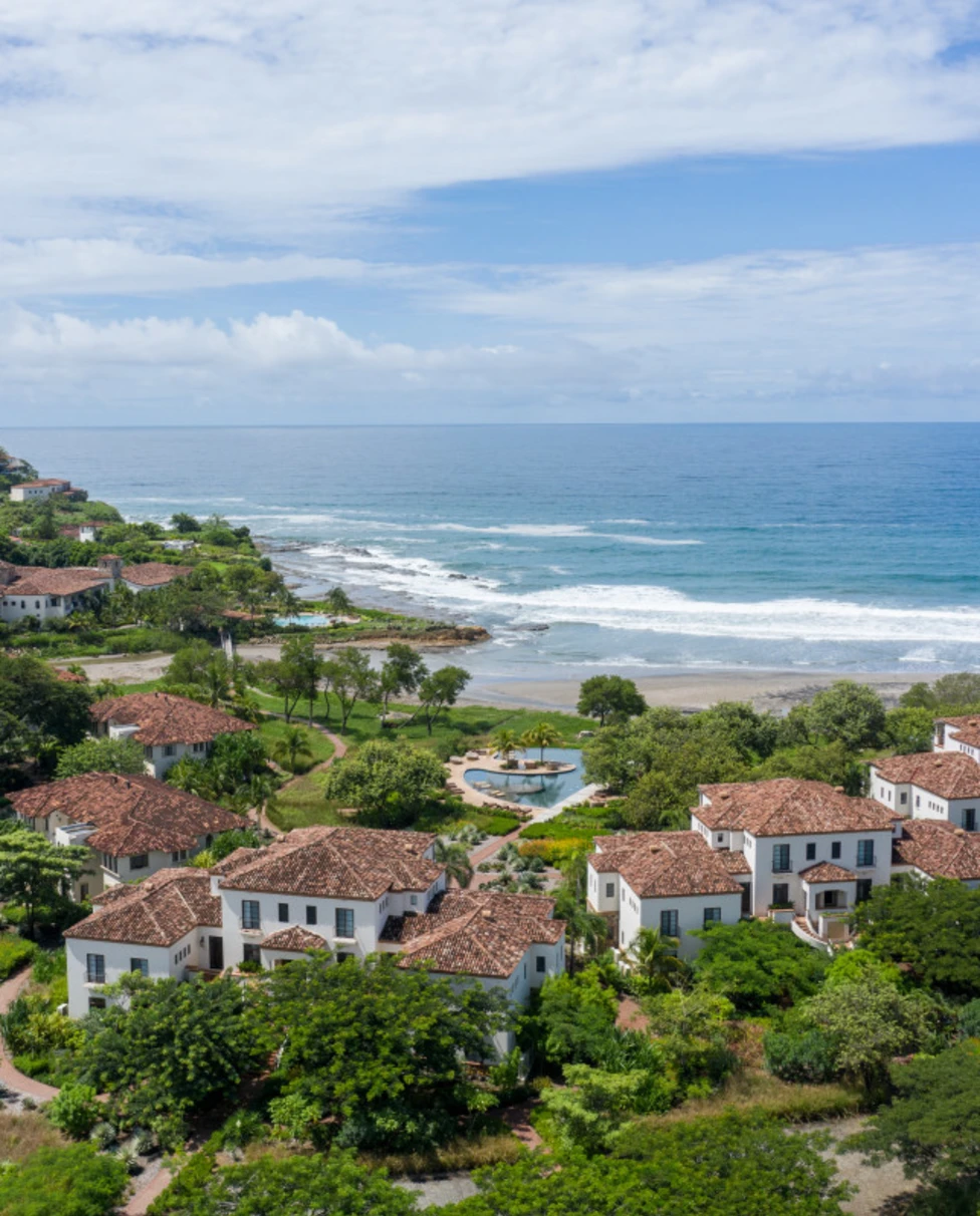A view of Rancho Santana with individual white buildings and tiled roofs surrounded by lush greenery and close to the ocean.