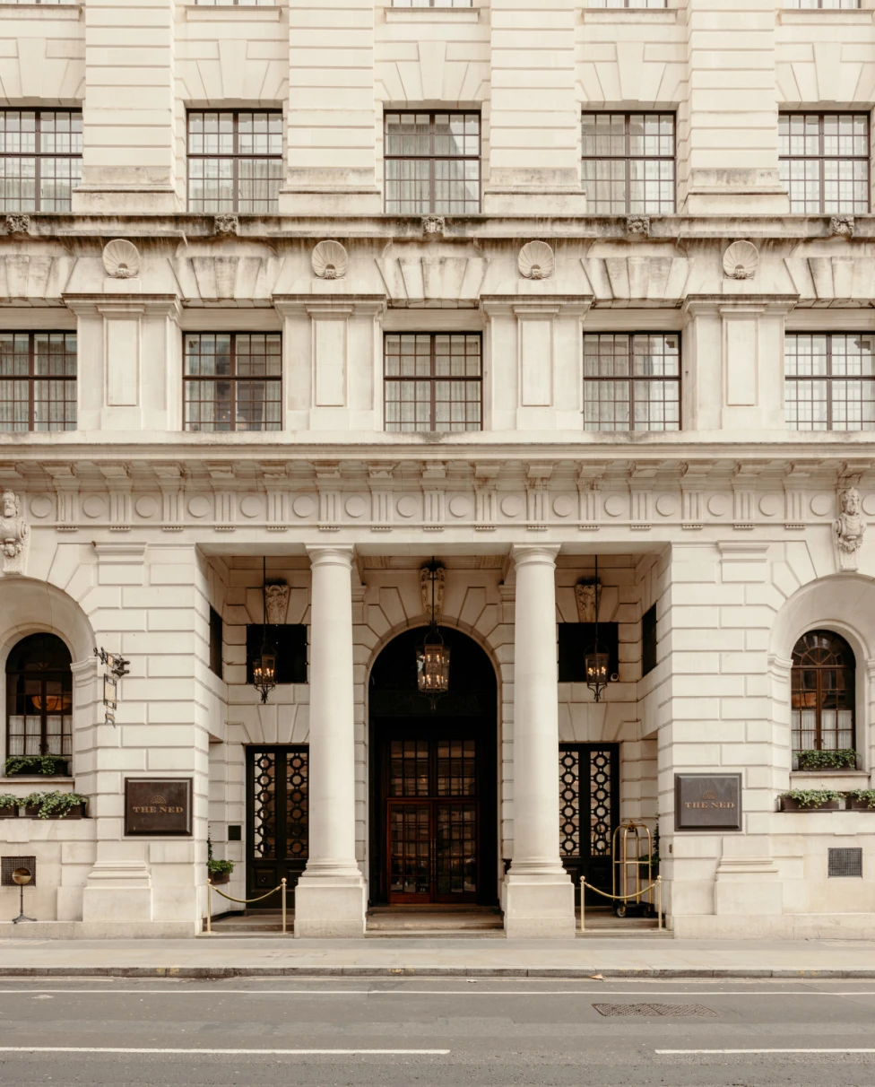 Elegant white facade of The Ned City of London with brown arched doorway and pillars.