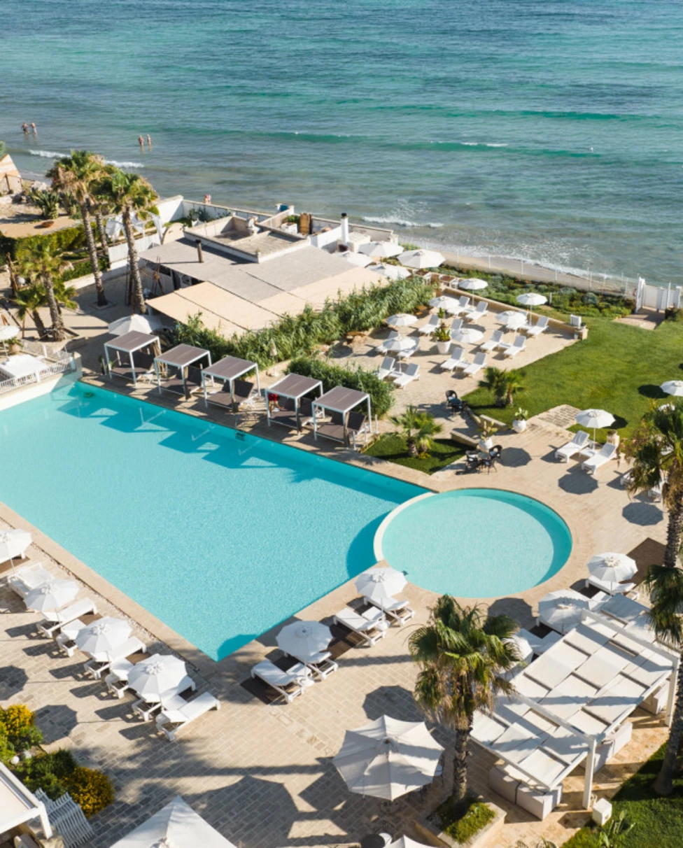 An aerial view of Canne Bianche hotel with a large pool area, sun loungers and grassy area by ocean.