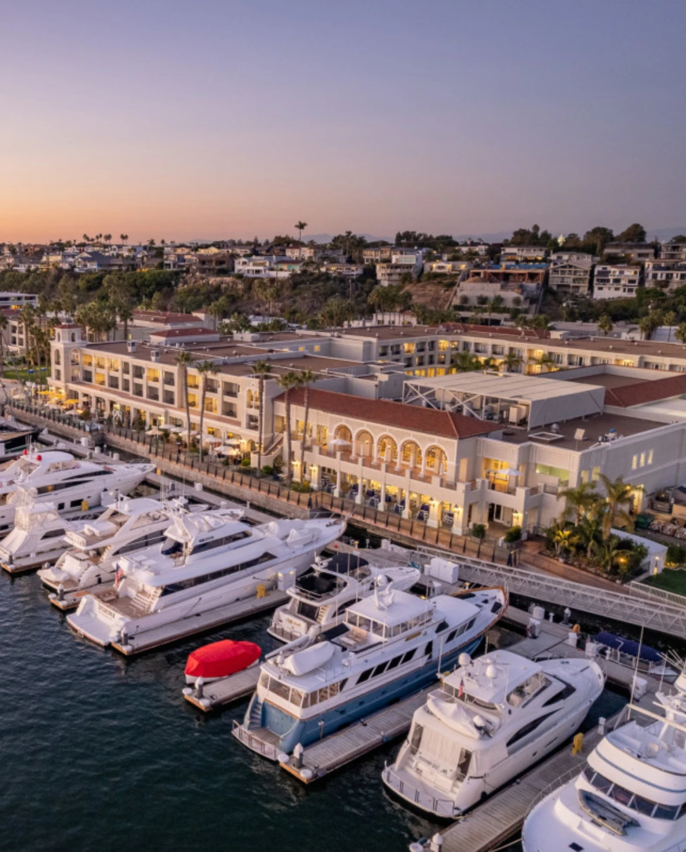 An aerial view of Newport Bay with many yachts docked outside the grand Balboa Bay Resort, taken in the evening at sunset.