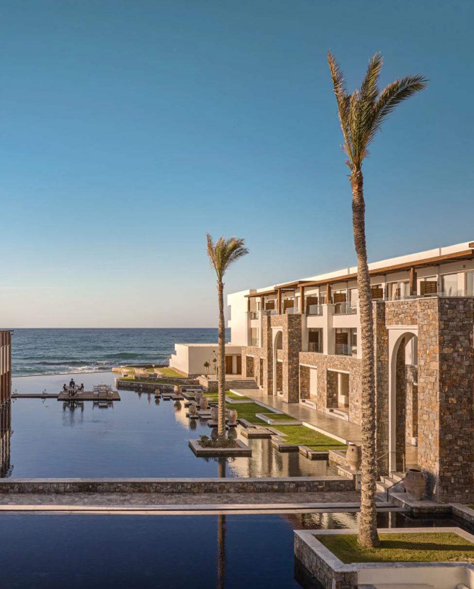 A view of the Amirandes hotel with pools cascading down the middle of the resort under palm trees, leading down to the ocean.