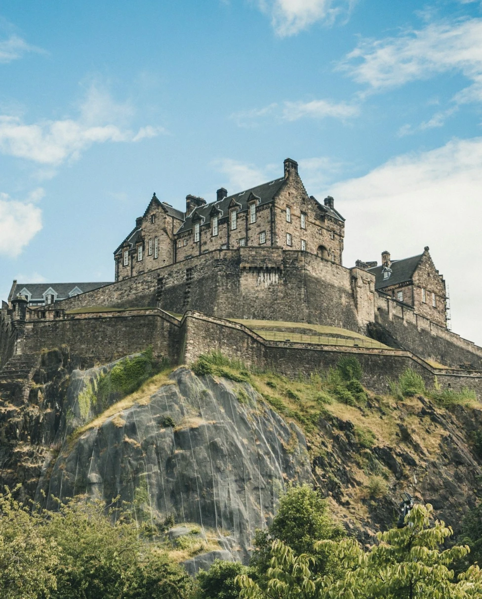 Edinburgh Castle, a historic fortress perched atop Castle Rock in Edinburgh, sits atop a rocky hillside on a sunny day dotted with clouds.