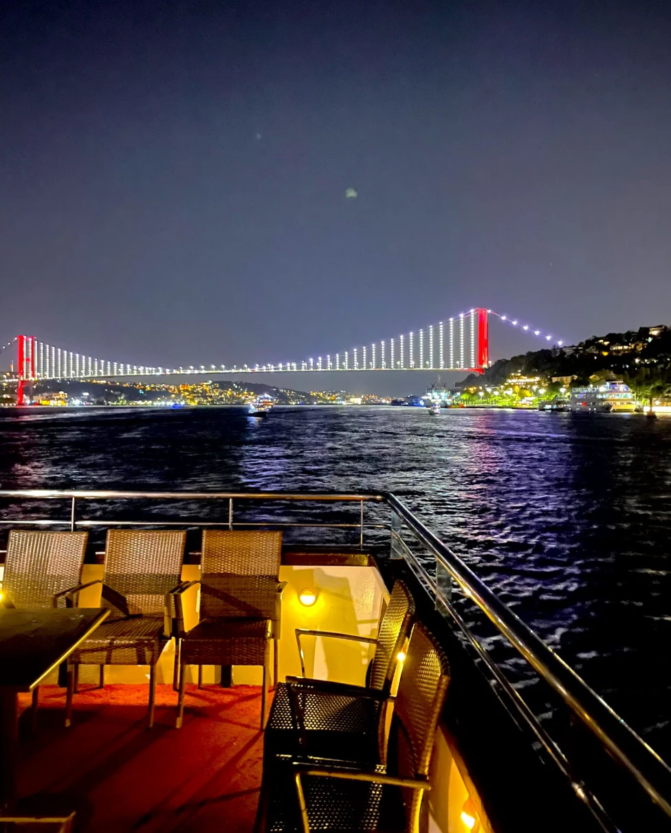A beautiful view of chairs on a balcony overlooking a river and colorfully arched bridge in the distance at nighttime.