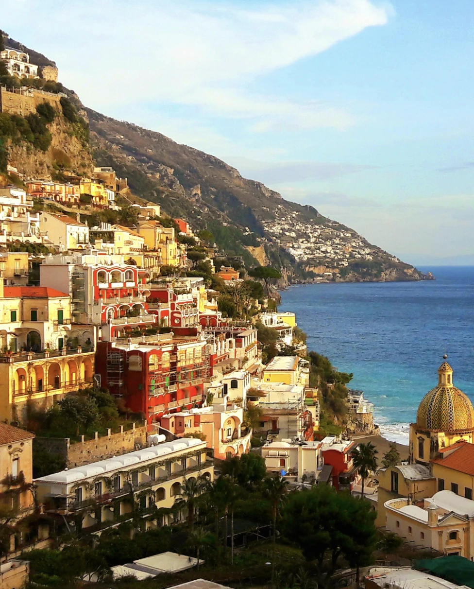A picture of the vibrantly colorful town of Positano, with various buildings nestled into a mountain. There is also a view of the blue sea in the background towards the right of the image.