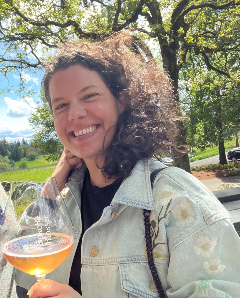 An individual holding a wine glass, with trees and a clear sky in the background.