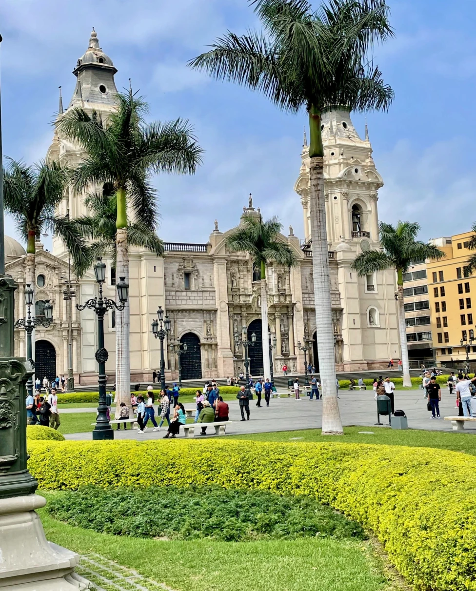 This image depicts a vibrant city square with a stone building, palm trees, a green hedge and people walking around in the surrounding areas.