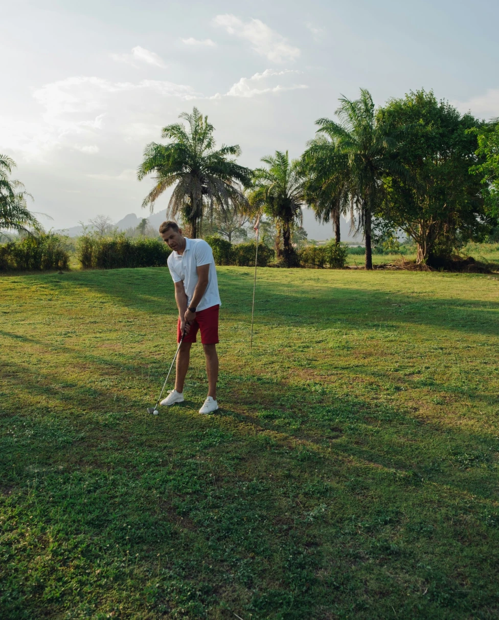 A golfer plays on a lush green course with trees and hills in the background.