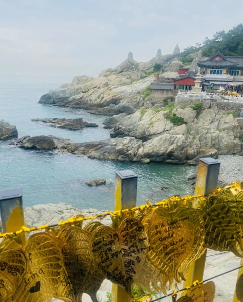 The image captures a rocky coastline with a structure and yellow ribbons in the foreground against hazy skies.