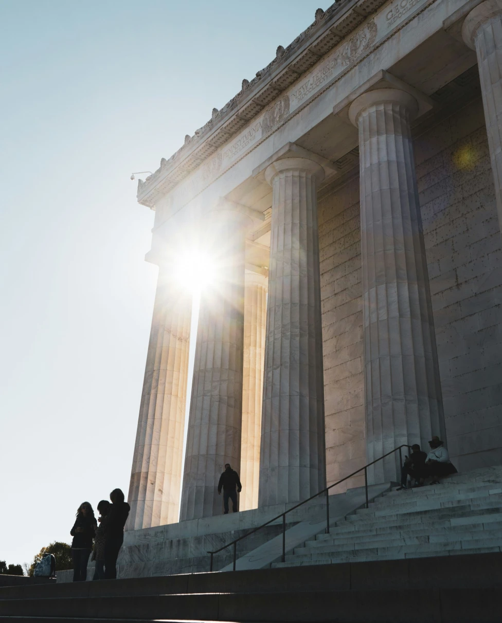 A picture of a white concrete building with pillars in front of it during daytime.