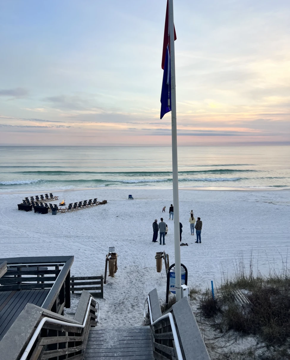 The image shows a serene white sand beach scene with people, a wooden boardwalk, a flag and chairs arranged in rows, set against a beautiful sunset or sunrise.