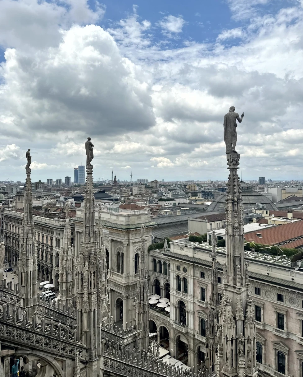 The image offers an aerial view of a cityscape with intricate gothic architecture in the foreground under a cloudy sky.