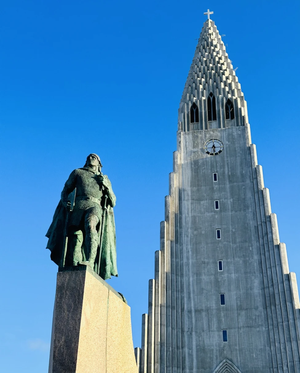 The image portrays a statue in front of a towering, pointed church against a backdrop of clear blue skies.
