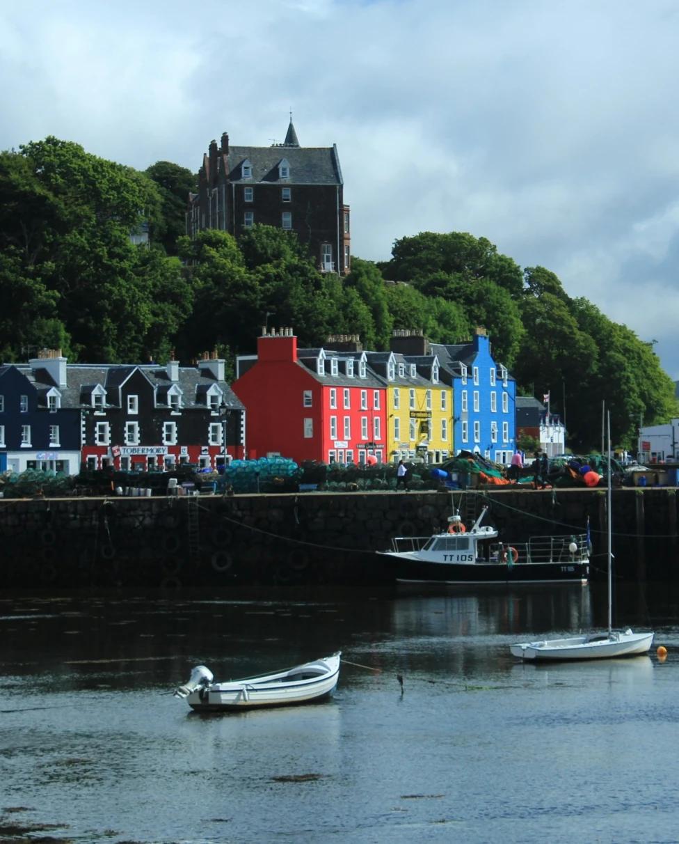 The image shows a scenic waterfront with colorful buildings, boats and a hill with greenery under a cloudy sky.