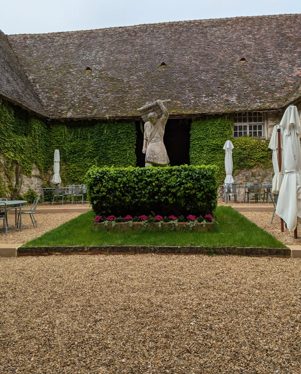 A serene garden courtyard with gravel ground, green hedges, sculptures and outdoor furniture.