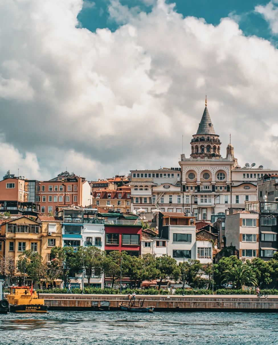 City view of Istanbul across a river with white boats and blue red peach black and white colored buildings, green trees, and a white clouds with a blue sky