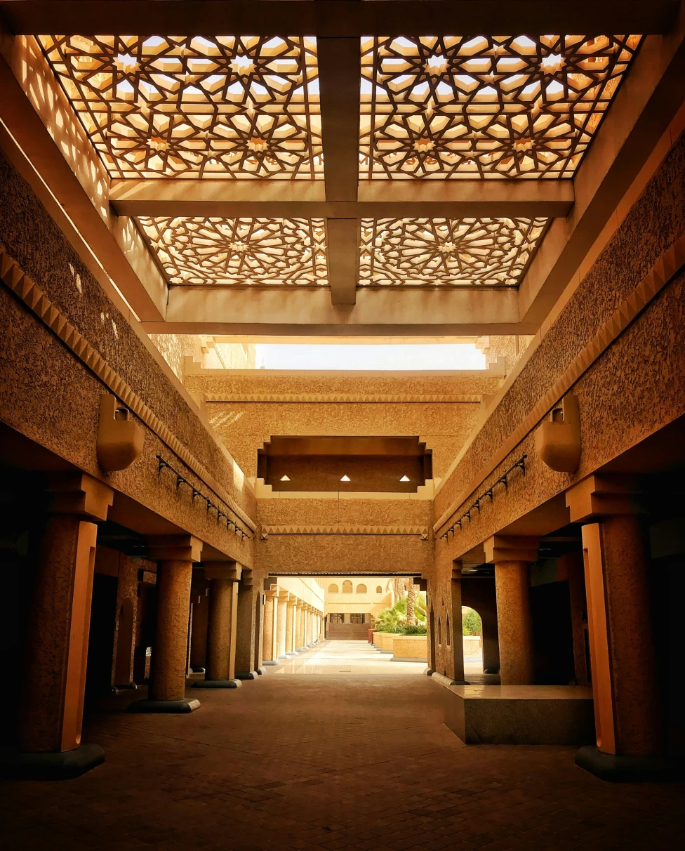 A picture taken inside of a corridor of a brown and white concrete building with a patterned roof.