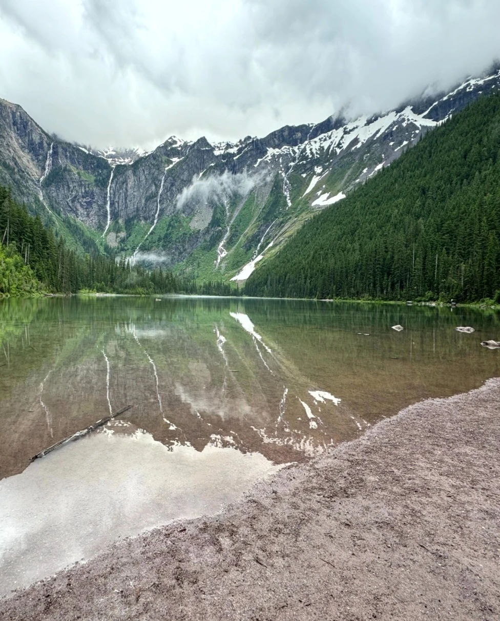The image captures a serene mountain lake with reflections of snow-capped peaks and forested slopes.