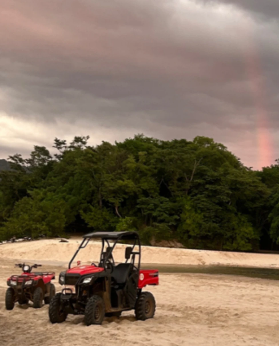 An ATV and UTV parked on a sandy shore with a faint rainbow over a forest under a cloudy sky.