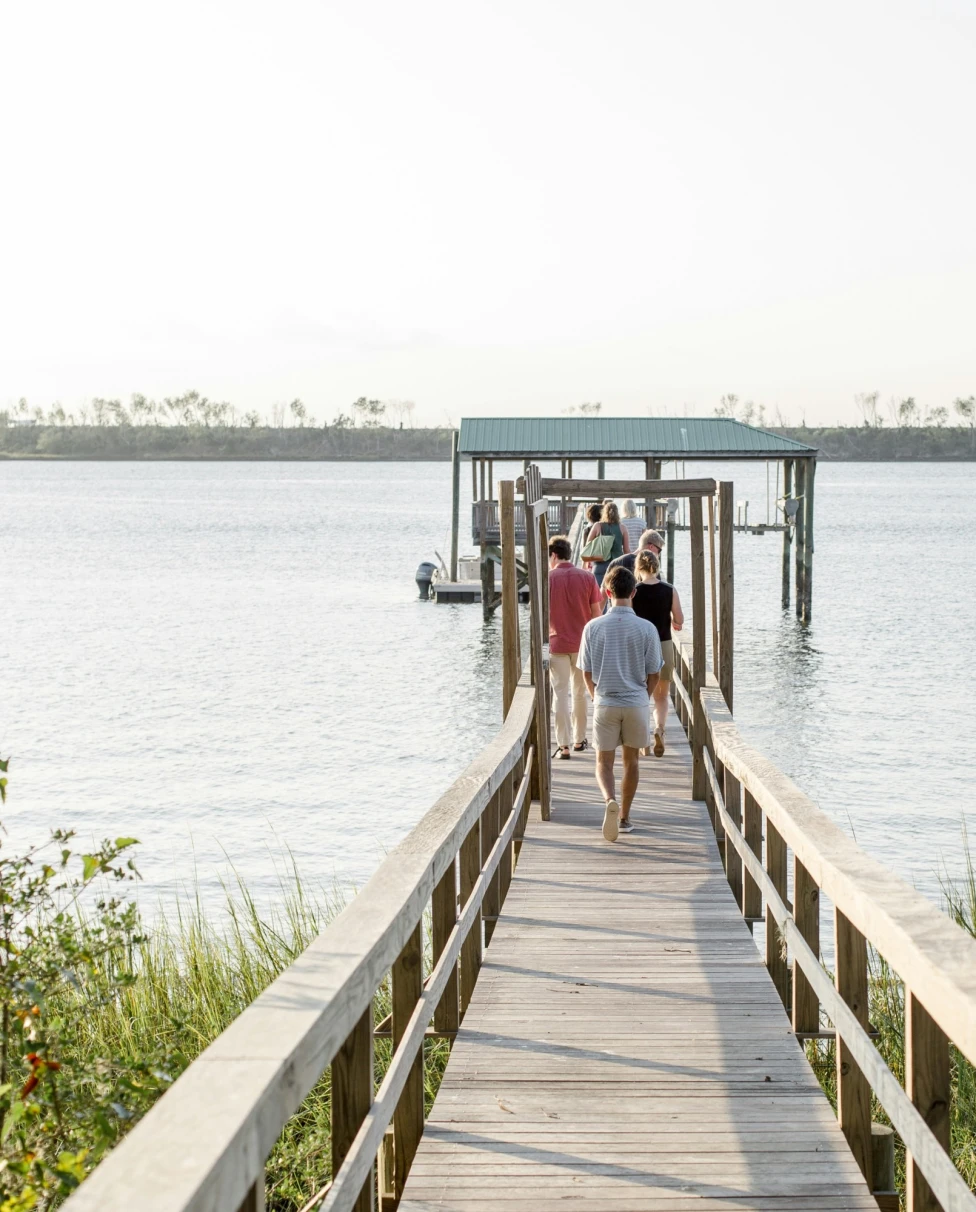 Three people are walking on a wooden pier towards a covered dock over a calm body of water, under a clear sky.